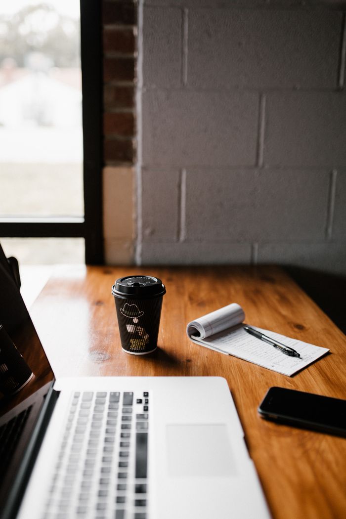 A cozy workspace featuring a laptop, coffee cup, notepad, and pen by a window.