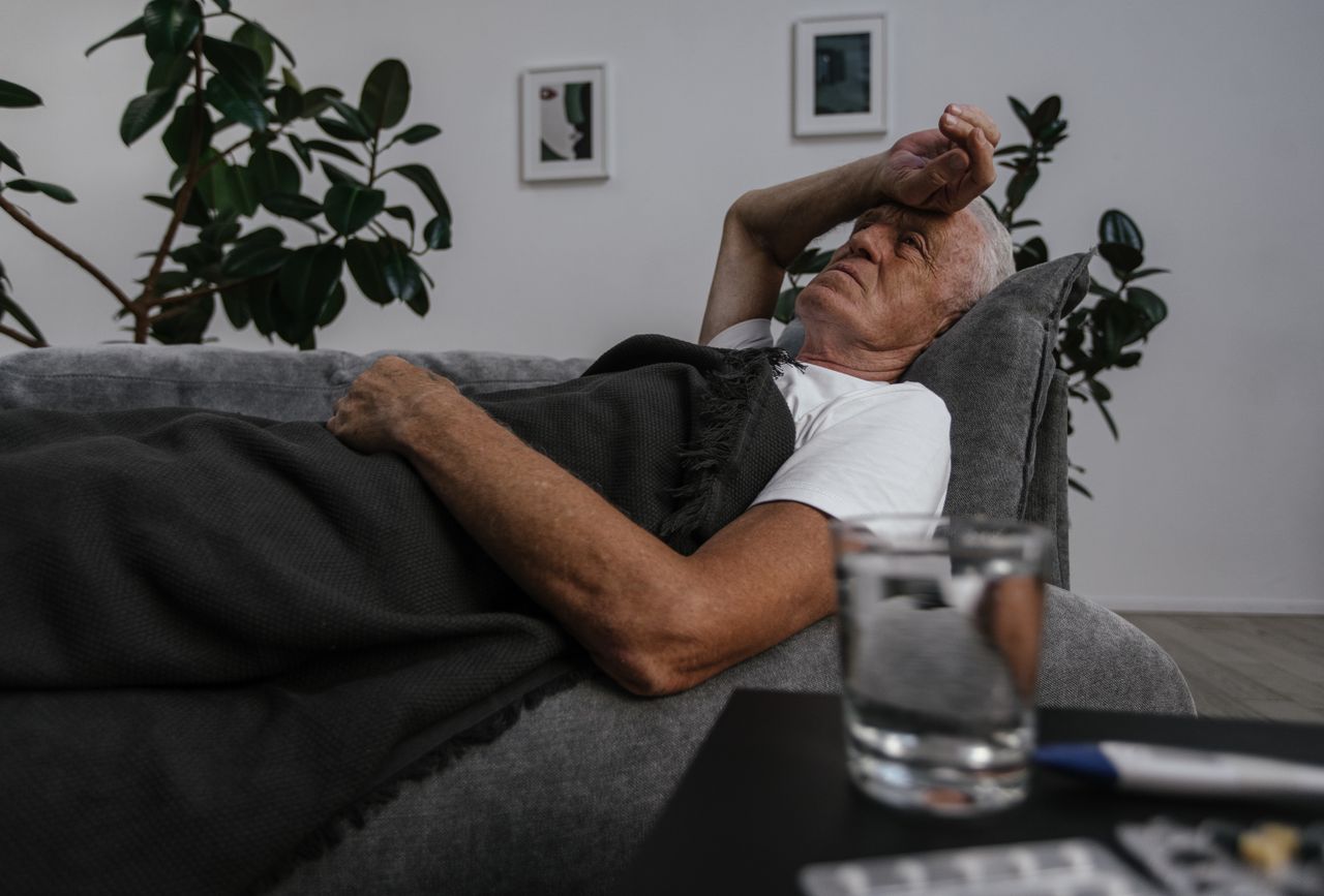 Elderly man lying on a couch with blanket, appearing unwell indoors.